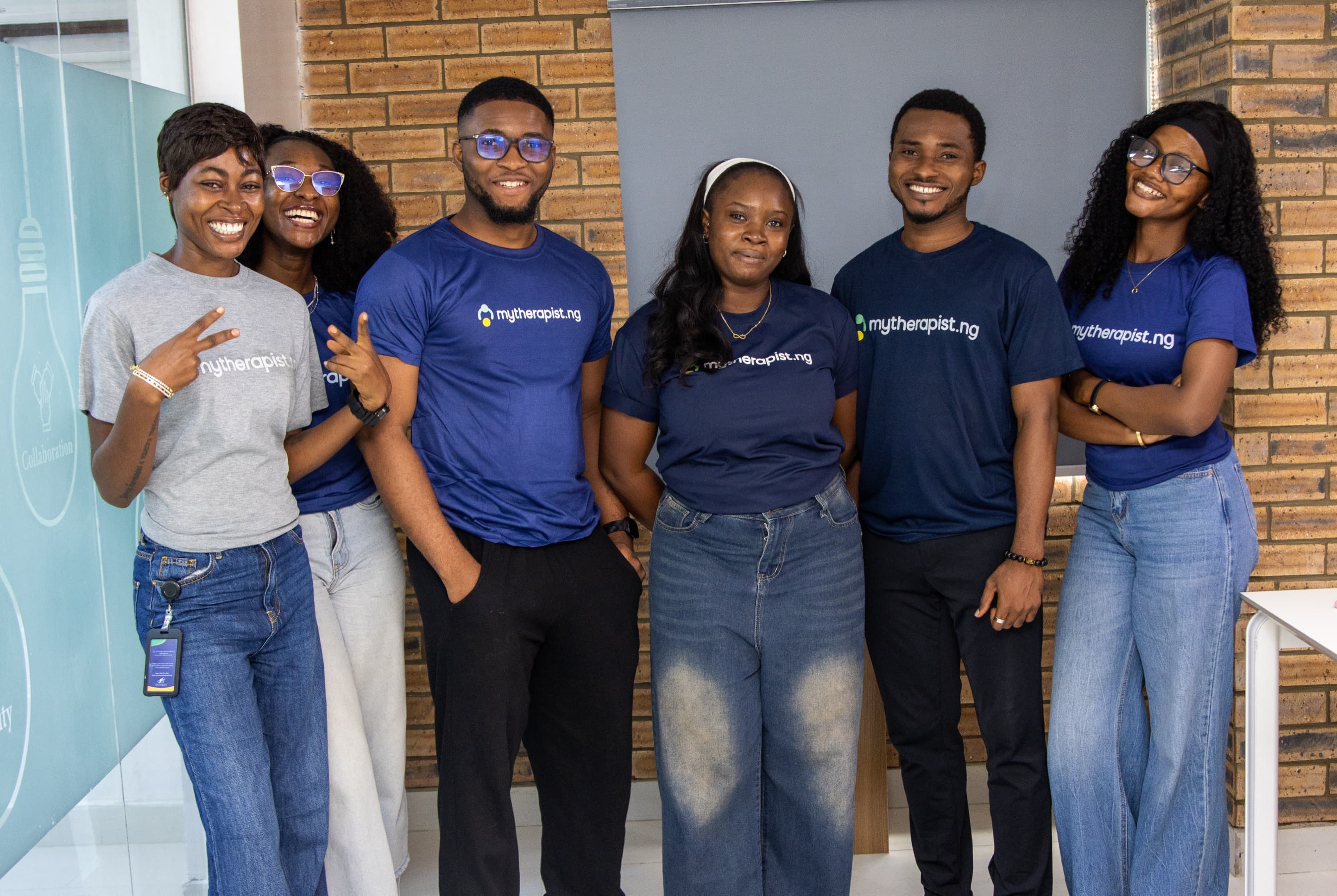 Six Mytherapist.ng team members standing together against a brick wall in the office, smiling
