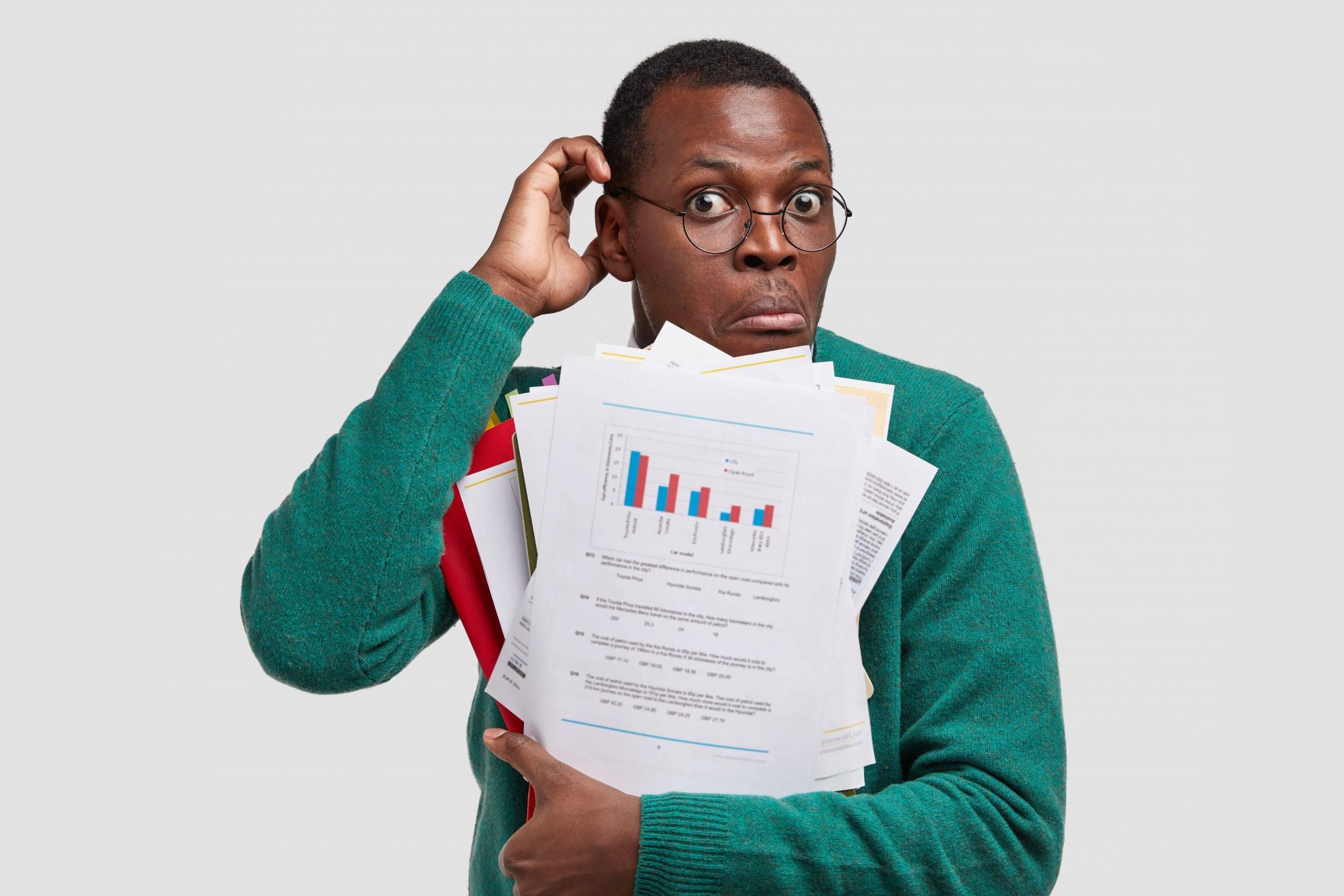 Indecisive black man scratches head, carries papers with data and diagram, feels puzzled as has to prepare financial report, isolated over white wall. Office employee with dark skin holds document