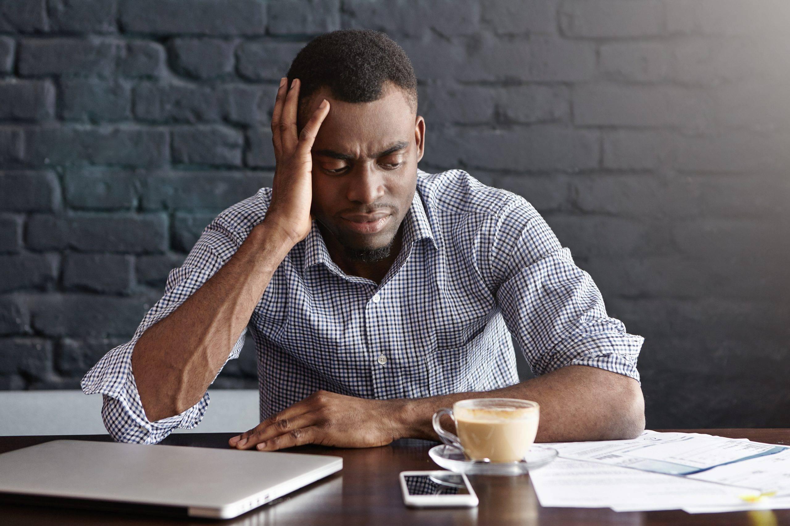 Signs your mental health is dwindling: Unhappy African businessman feeling stressed and frustrated, facing financial troubles, leaning elbow on table with mug, laptop, cell phone and papers while doing finances during coffee break