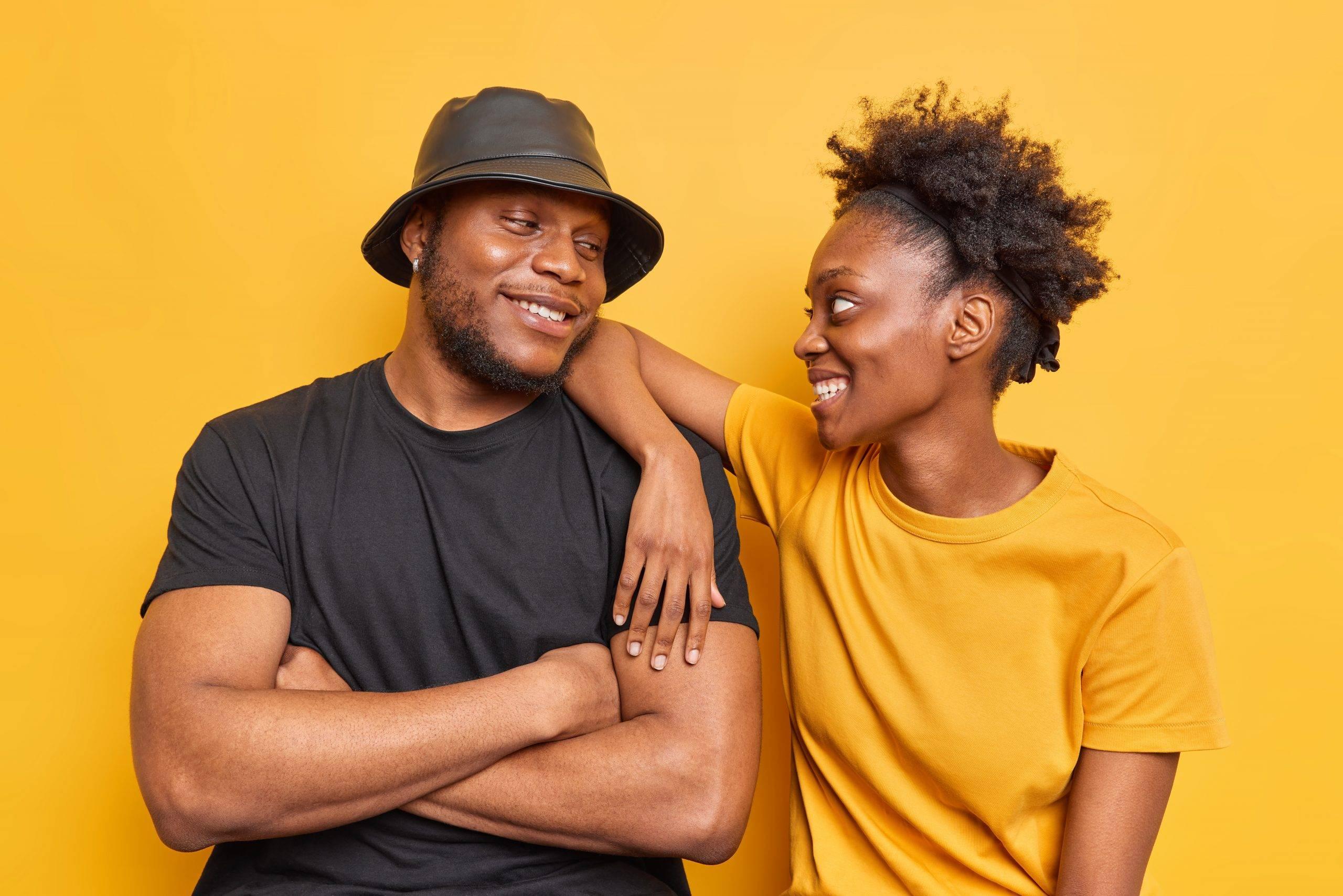 Positive dark skinned young woman and man have fun look gladfully at each other wear casual yellow and black t shirt pose indoor. Happy curly haired teenage girl leans at shoulder of best friend showing support, one of the best ways to help a friend with depression.